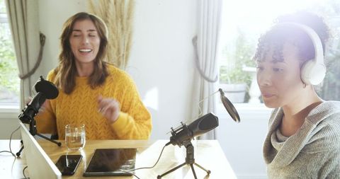 Female cohosts recording home podcast with professional microphones headphones tablet sunlit