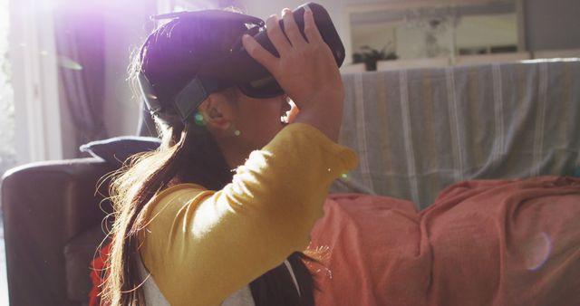 Girl using VR headset in sunlit living room, highlighting modern technology and home entertainment. Ideal for content on youth engagement with technology, gaming, futuristic experiences, or at-home leisure activities.