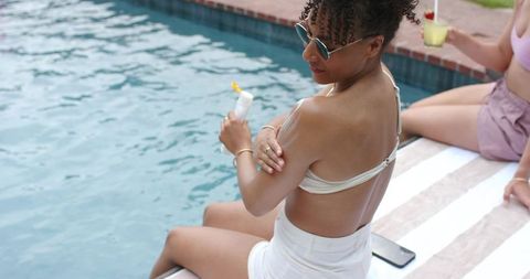Applying sunscreen poolside on striped towel with cocktail and smartphone nearby