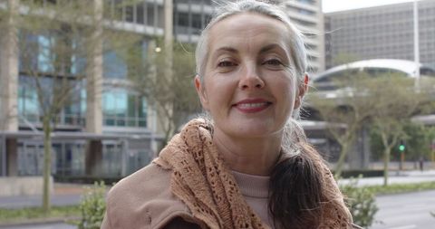 Mature woman smiling on urban sidewalk wearing tan knit scarf and light jacket, office backdrop