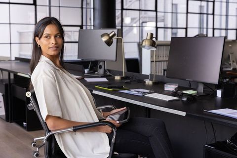 Professional woman turning toward computer monitors while holding tablet in modern office