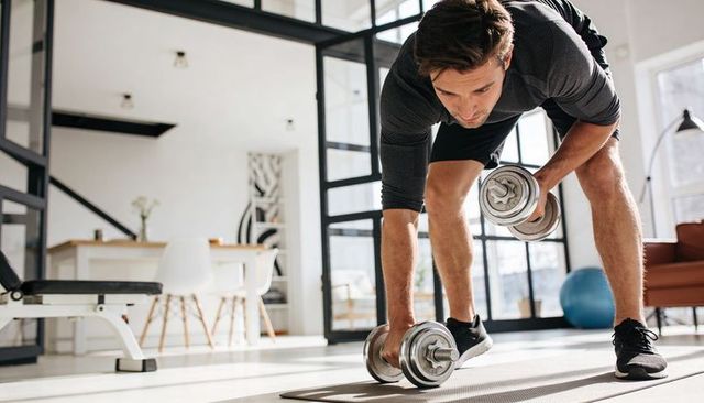 Fit man performing dumbbell row at home gym in bright modern living space for workout