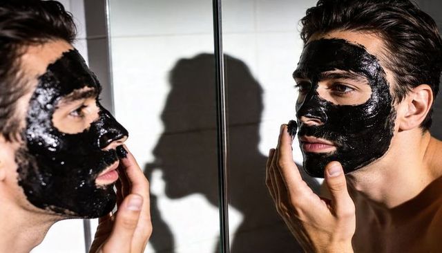 Bare-chested man applying charcoal peel-off face mask in bathroom mirror