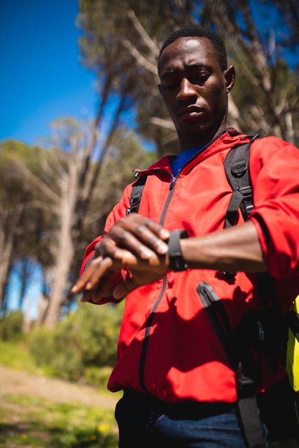 African American man checking his smartwatch while trekking in a forest. He is wearing a red jacket and carrying a backpack, indicating an adventure or travel activity. This image can be used for promoting outdoor activities, fitness technology, travel adventures, and active lifestyles.