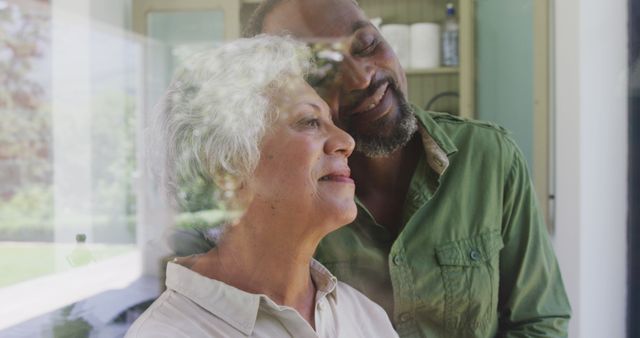 Happy senior biracial couple standing by window embracing and smiling. Love, senior lifestyle, togetherness and domestic life.