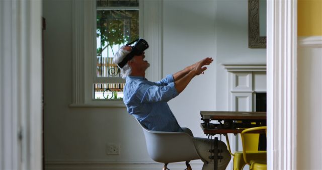 Senior man enthusiastically using virtual reality headset while sitting in a stylish home office with white walls and contemporary furnishings. Ideal for showcasing innovations in technology for seniors, promoting VR experiences, or highlighting modern tech-savvy lifestyles among the elderly.
