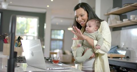 Mother using laptop in kitchen while holding baby