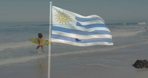 Waving uruguay flag on sunny beach with surfer carrying yellow surfboard by shoreline