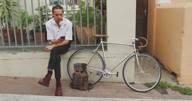 Young man in an urban environment sitting on a sidewalk with his bicycle. He is casually dressed, wearing a striped shirt, black jeans, and red shoes, holding and looking at his smartphone. A backpack lies next to him. Suitable for illustrating modern lifestyle, travel, commuting, and leisure activities.