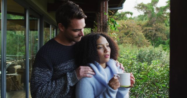 Young couple on a balcony, warmly dressed, enjoying a peaceful morning. The woman is holding a coffee cup while the man gently embraces her. Use this image for themes related to romance, relaxation, morning routines, and nature retreats.