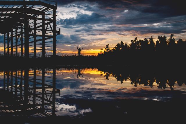 View of an unfinished building structure silhouetted against a vibrant sunset sky with colorful clouds. The still waters reflect the scene, adding tranquility and beauty. Ideal for using in contexts illustrating construction, nature, scenic beauty, and peaceful evening atmospheres.