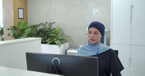 Muslim Receptionist Wearing Hijab Working at Modern Reception Desk with Computer and Plants