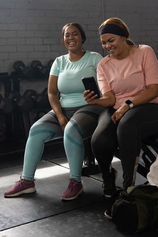 African american women laughing and checking phone while resting on gym bench