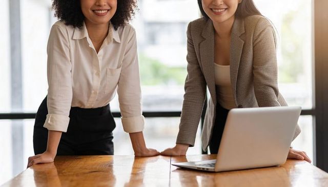 Female professionals leaning over wooden table in bright office with laptop and smiles