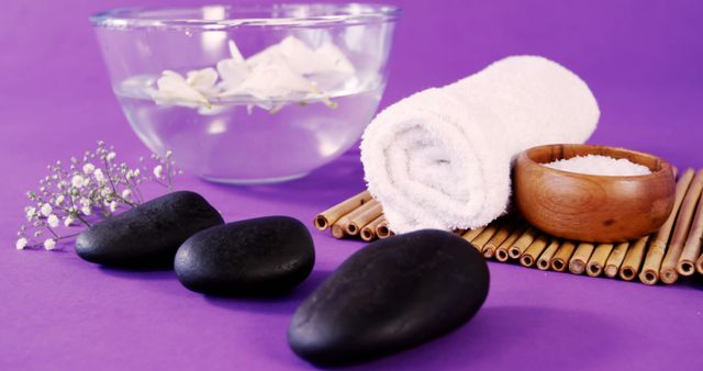 A serene spa setup features smooth black stones, a white towel, and a bowl of water with flower petals, with copy space. Elements like the bamboo mat and lavender sprigs add to the tranquil and rejuvenating atmosphere of the scene.