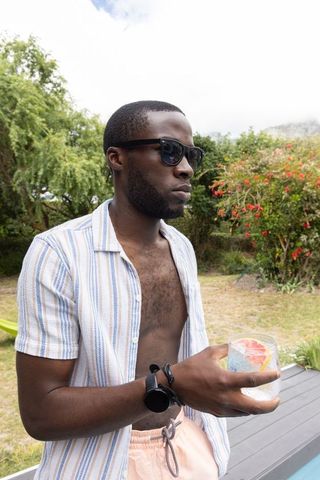 Relaxed african american man standing on pool deck wearing sunglasses holding citrus drink