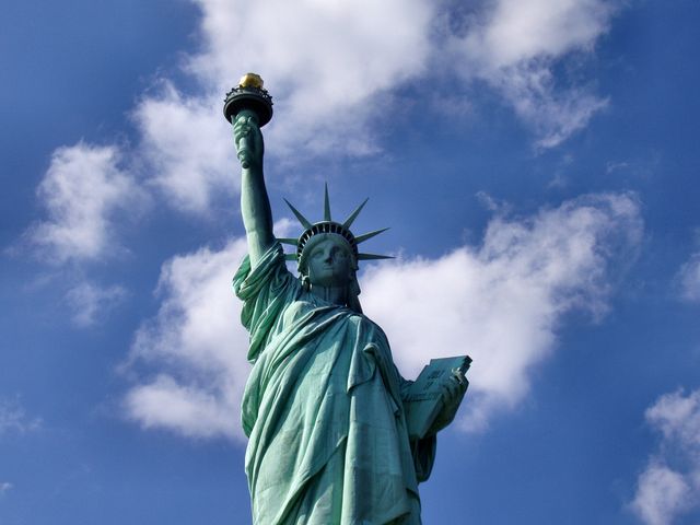 Statue of Liberty towering against a bright blue sky with scattered white clouds. Perfect for travel publications, educational materials, promotional content on tourism in New York City, and patriotism-themed editorials.
