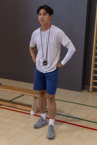 Focused asian male coach standing in gym holding stopwatch hands on hips athlete training