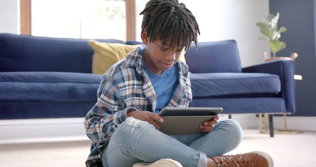 African American boy sitting on floor in living room, wearing casual clothes, and using tablet for online learning. Blue couch and indoor plant in background suggest cozy home environment. Suitable for illustrating topics related to online education, technology in learning, homeschooling, and modern childhood activities.