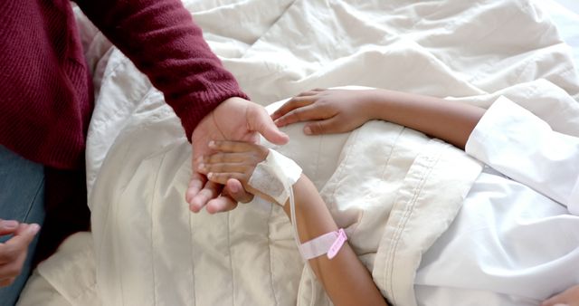 Midsection of african american girl patient and her mother holding hands in bed in hospital room. Medicine, healthcare and hospital.