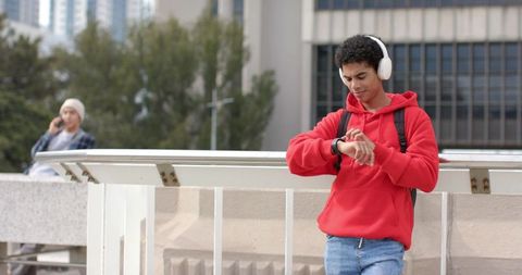 Young man in red hoodie checking smartwatch while listening to music on urban overpass
