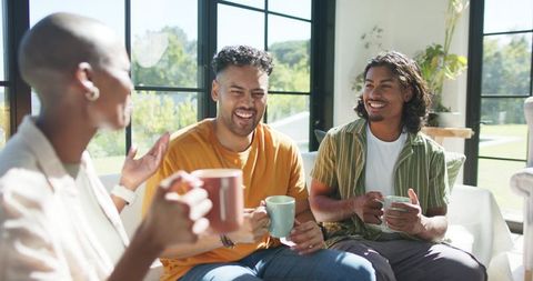 African american friends laughing sipping coffee and chatting in sunlit modern living room