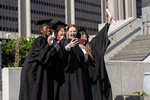 Multicultural female graduates celebrating selfie on campus with diplomas and mortarboards