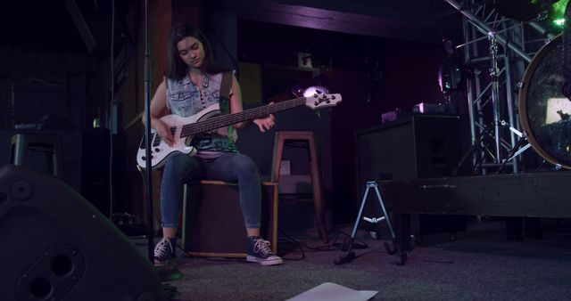 Young female musician practicing bass guitar on stage at a music venue, possibly preparing for a live performance or rehearsal. Useful for advertising music events, showcasing musical talent, or promoting musical instruments and equipment.