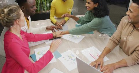 Diverse creative team stacking hands over charts and laptop, collaborative office energy