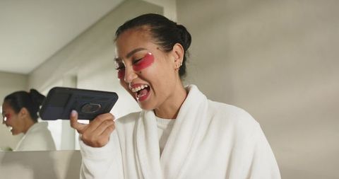 Mid-adult asian woman laughing while wearing robe and red eye patches holding smartphone at vanity