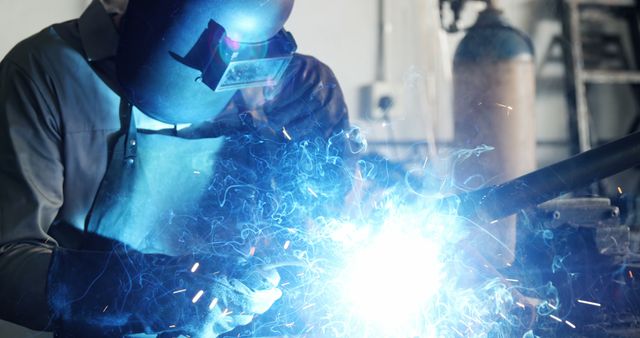 Person wearing protective welding helmet and gear working with bright light and sparks coming from welding equipment. Useful for illustrating industrial work environments, safety practices, skilled trades, and manufacturing jobs in various publications and educational materials.