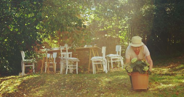 Woman wearing a straw hat bends over to pick up large potted plant in a sunlit garden area. Background features a rustic wooden table and chairs set up under shade, suggesting an outdoor dining area. Suitable for use in lifestyle blogs, gardening websites, or summer relaxation promotions.
