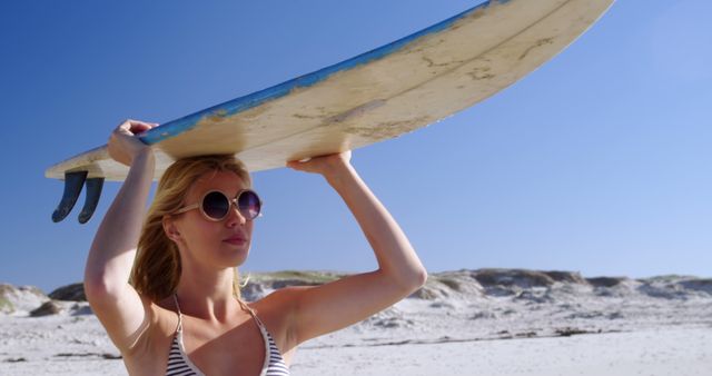 A young Caucasian woman carries a surfboard on a sunny beach, with copy space. Her relaxed demeanor and stylish sunglasses suggest a leisurely day of surfing ahead.