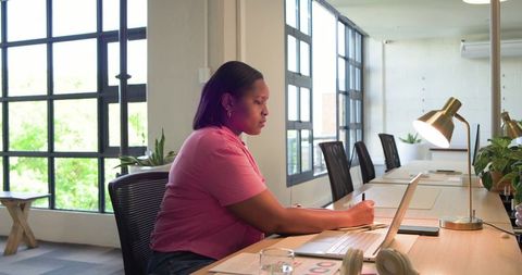 African american woman working on laptop in coworking space with plants and brass lamp