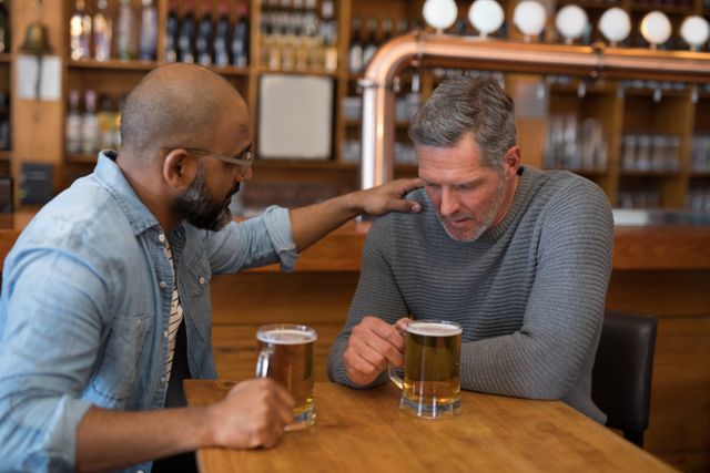 Two men sitting at a bar, one comforting the other who appears to be upset. Both have beers in front of them. Ideal for themes of friendship, emotional support, mental health awareness, and social interactions. Can be used in articles, blogs, or advertisements focusing on men's mental health, support systems, and the importance of friendship.