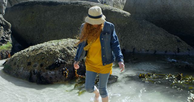 This stock image features a young woman with red hair walking along a rocky beach. She is wearing casual attire, including a denim jacket, jeans, and a straw hat. The woman appears to be enjoying the warm summer weather as she explores the natural surroundings. This image is perfect for use in travel blogs, summer vacation promotions, fashion websites emphasizing casual wear, or outdoor adventure advertisements.