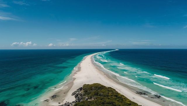 Aerial view of narrow white sand spit curving into turquoise ocean with footprints and distant figur