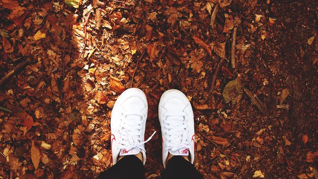 Person wearing white sneakers standing on forest floor covered with dry autumn leaves. Warm and earthy tones suggesting a seasonal backdrop suitable for promoting outdoor activities, fall fashion, nature exploration, or leisure time in nature.
