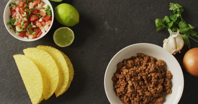 A top-down view of taco ingredients laid out on a dark countertop. Elements include crispy taco shells, a bowl of seasoned ground beef, chopped salsa mix in a small bowl, and fresh ingredients such as lime, onion, garlic, and cilantro. Perfect for use in recipes, cooking blogs, Mexican cuisine promotions, and food preparation magazines.