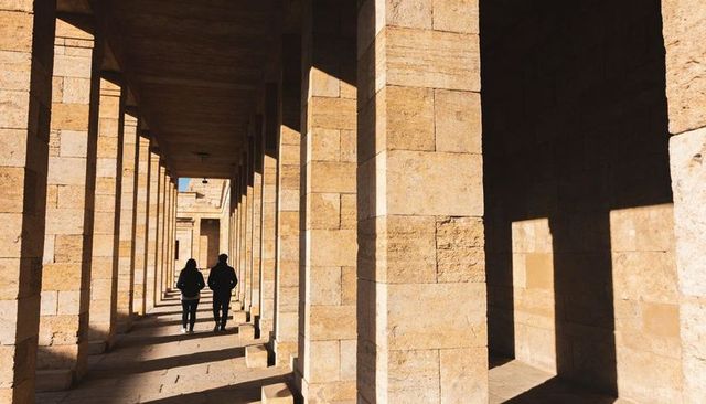 Two Adults Walking Through Sunlit Sandstone Colonnade Casting Strong Geometric Shadows