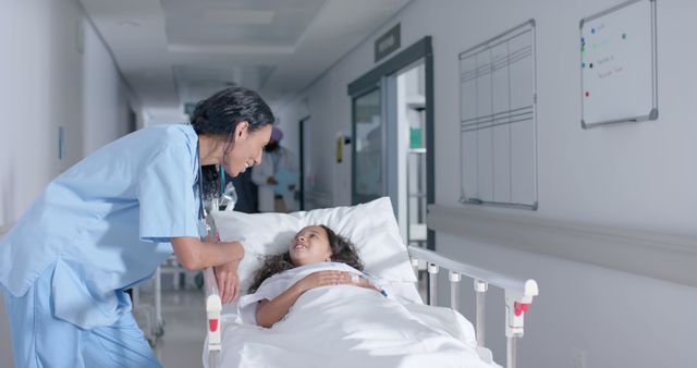 Nurse leaning over to interact with young patient lying in a hospital bed in a well-lit hospital corridor. This image may be used for healthcare promotions, websites about medical care, insurance advertisements, or articles discussing hospital staff and patient interactions.