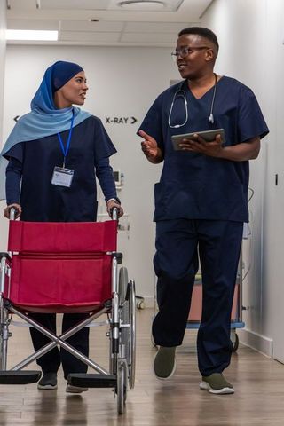 Multicultural medical team walking corridor with wheelchair and tablet in radiology