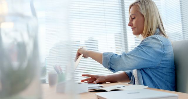 Blond woman in casual blue denim shirt sitting at desk, studying with notebooks and pen in bright office setting. This image can be used for educational materials, work productivity blogs, articles on women's empowerment, business presentations, advertisements for education services, or home office inspiration.