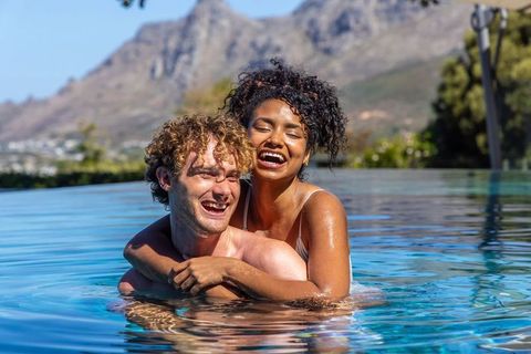 Diverse couple laughing and hugging in infinity pool at sunny mountain resort relaxing