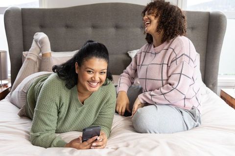 African american friends laughing and using phones while lounging on bed in cozy bedroom
