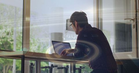 Focused Young Man Typing on Laptop Near Window with Digital Overlay