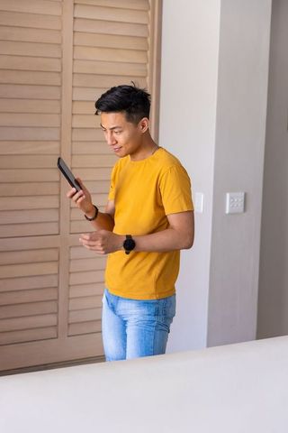 Asian man standing with tablet near louvered doors wearing mustard tee and jeans