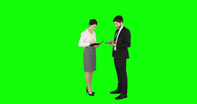 Business professionals in stylish office attire are engaging with digital devices while standing against a green screen background. This can be used for various purposes, including creating promotional materials, presentations, or educational videos requiring a customizable backdrop. The green screen allows for easy background removal and replacement, making the scene versatile for different business and marketing contexts.