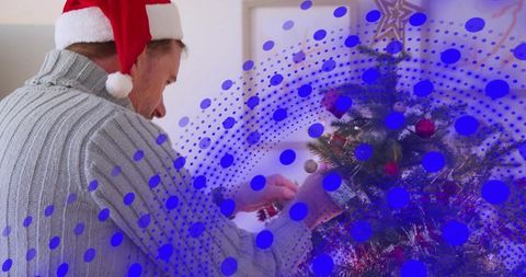 Man wearing santa hat decorating christmas tree with ornament, blue halftone overlay
