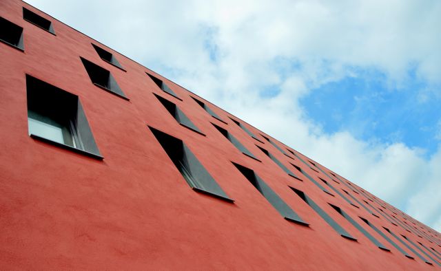 Modern architectural building with distinct red façade featuring asymmetrical windows. Blue sky with scattered clouds in the background enhances the visually striking design. Ideal for urban architecture, design concepts, and modern construction themes in advertisements or editorial content.